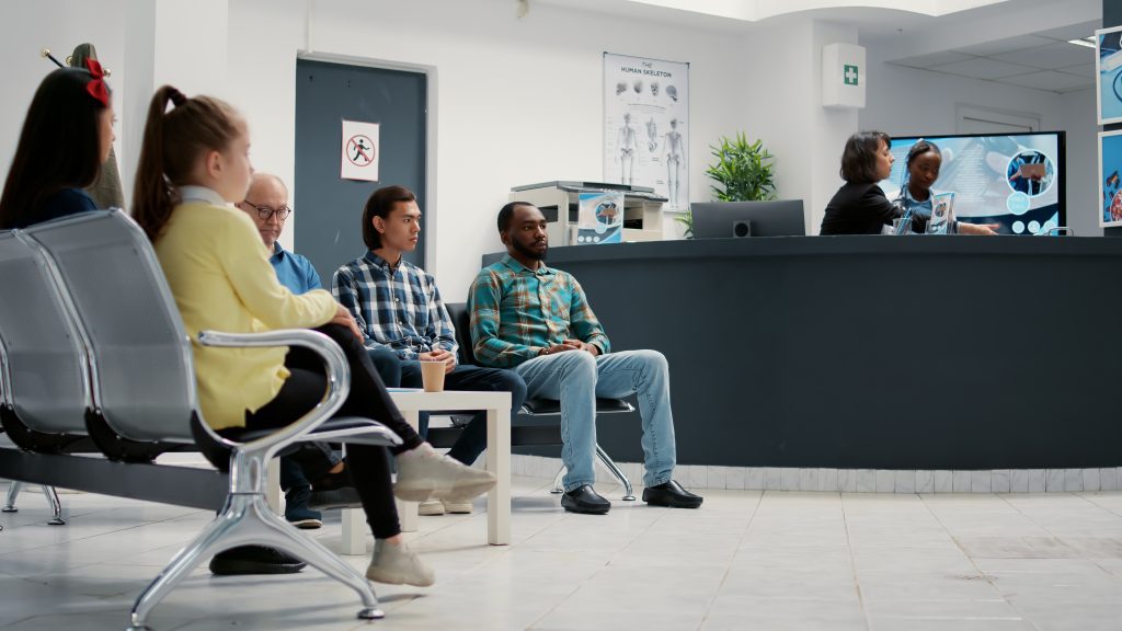 Grupo diverso de pessoas esperando na recepção do hospital para a consulta médica com o clínico geral. Pacientes na sala de espera sentados na clínica de saúde. Plano da câmera em tripé. Intuito de otimizar o tempo de espera dos pacientes.