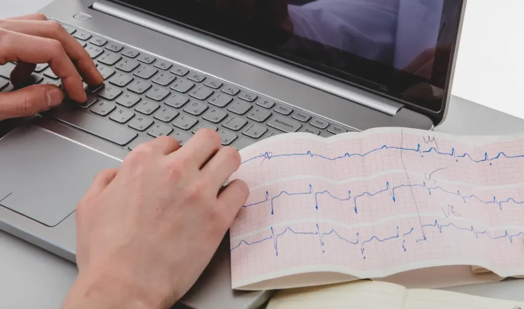 Imagem de uma pessoa usando um notebook, com um eletrocardiograma impresso ao lado.