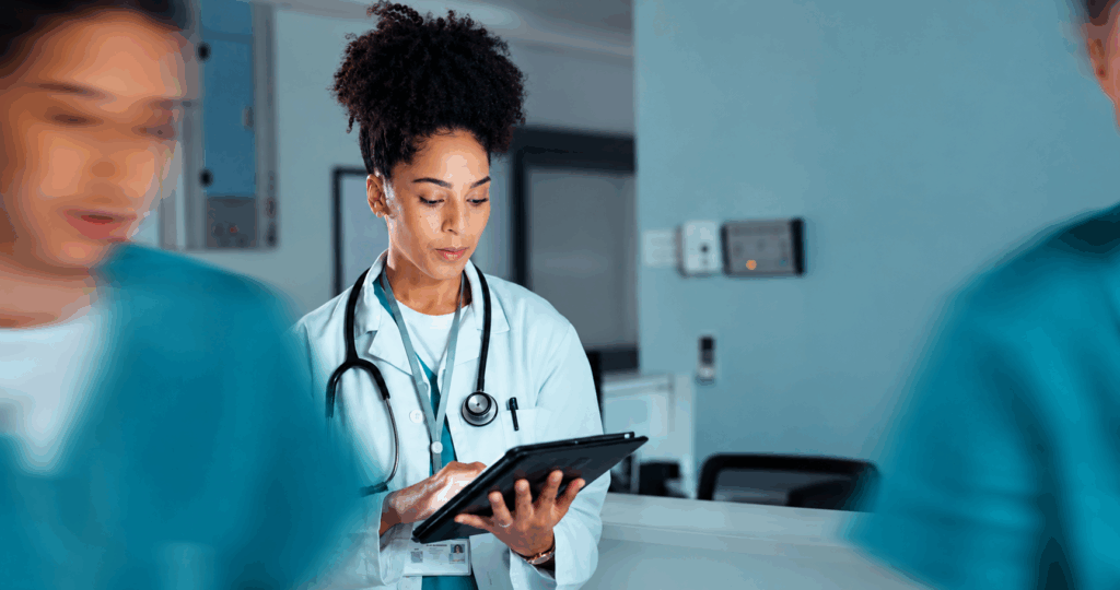 Profissional de saúde negra, usando jaleco branco e estetoscópio, consulta um tablet em ambiente hospitalar, enquanto outros profissionais aparecem desfocados ao fundo e nas laterais.